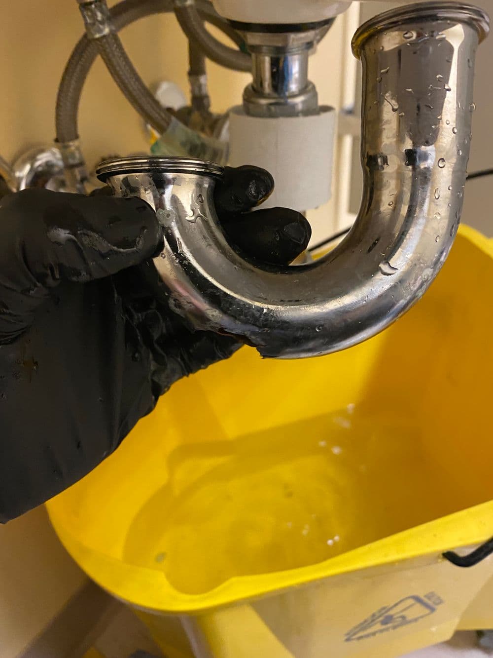 Person in a black glove holding a shiny plumbing trap above a yellow bucket with water.