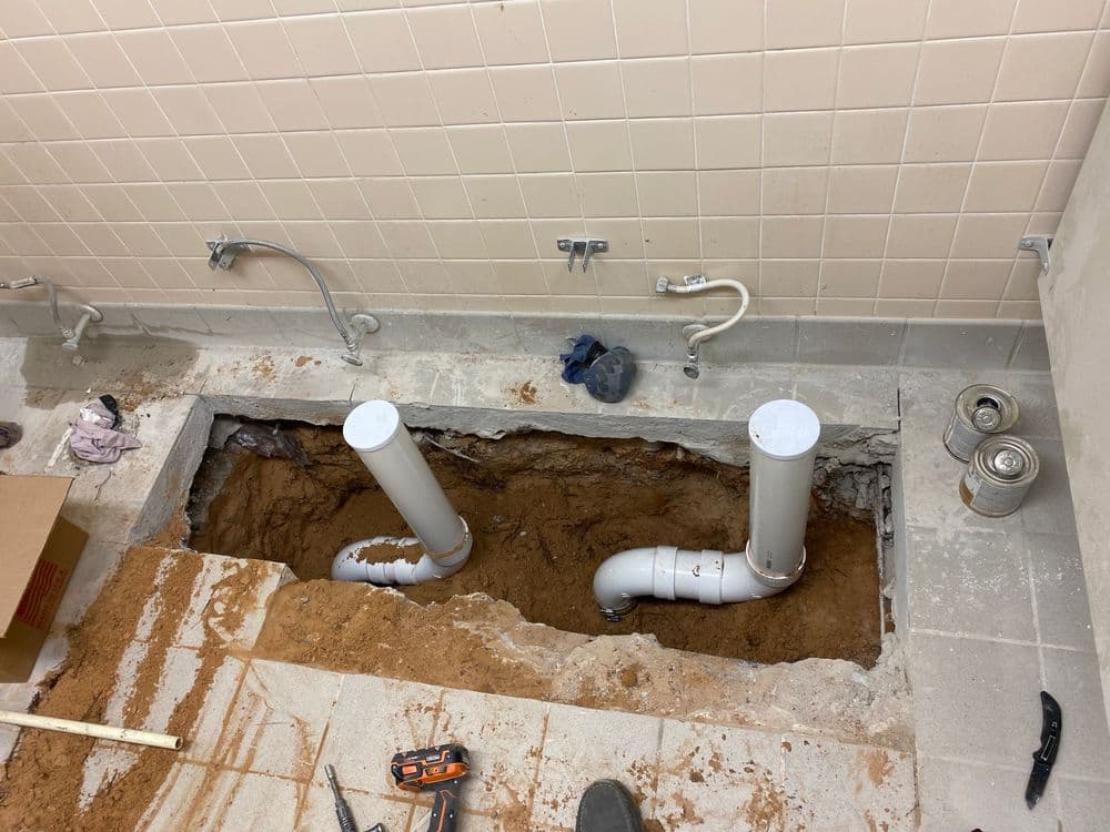 Plumbing repair in progress with exposed pipes and trench in a bathroom floor.