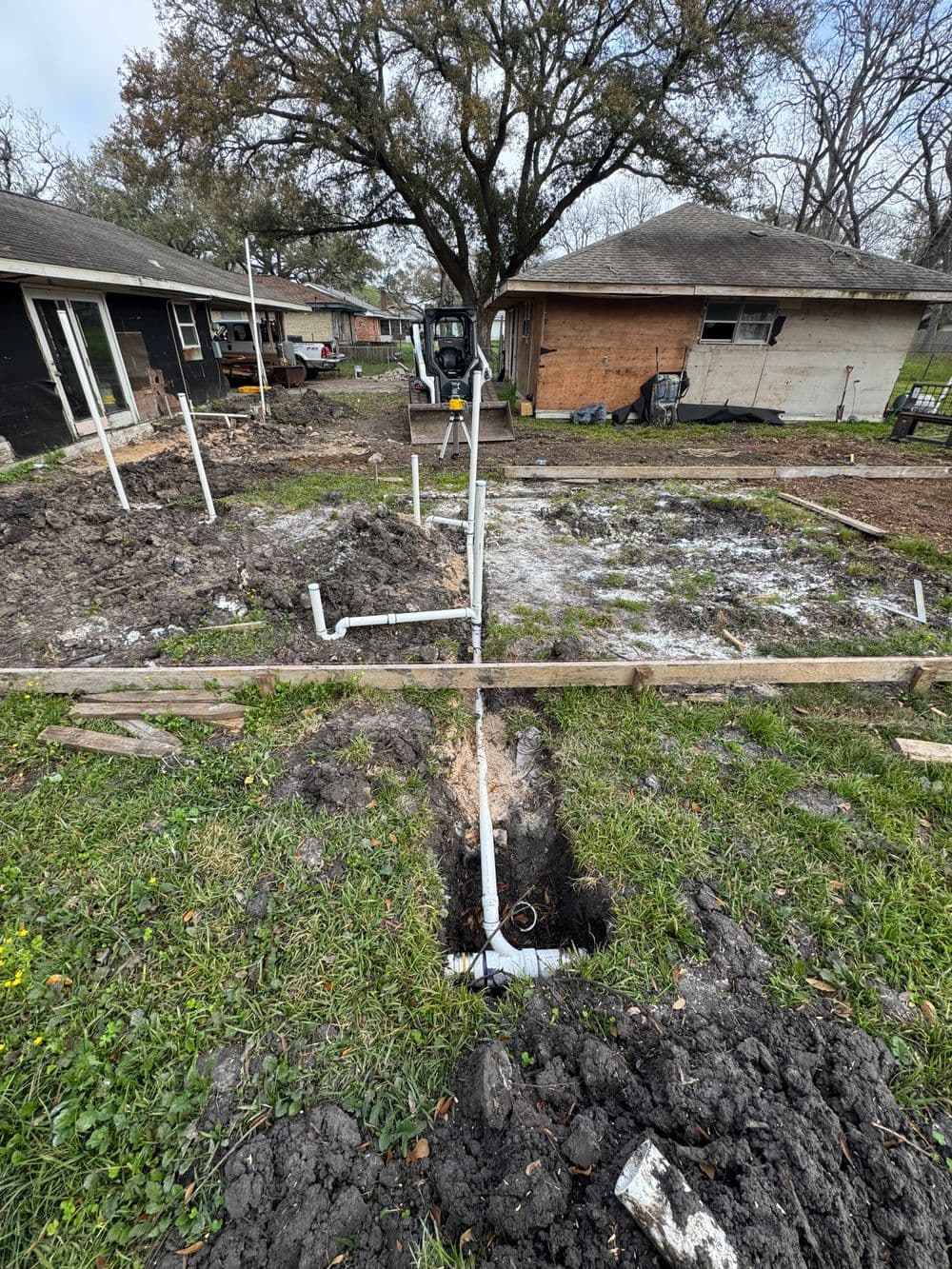 Construction site with excavated foundation, visible plumbing pipes, and surrounding vegetation.