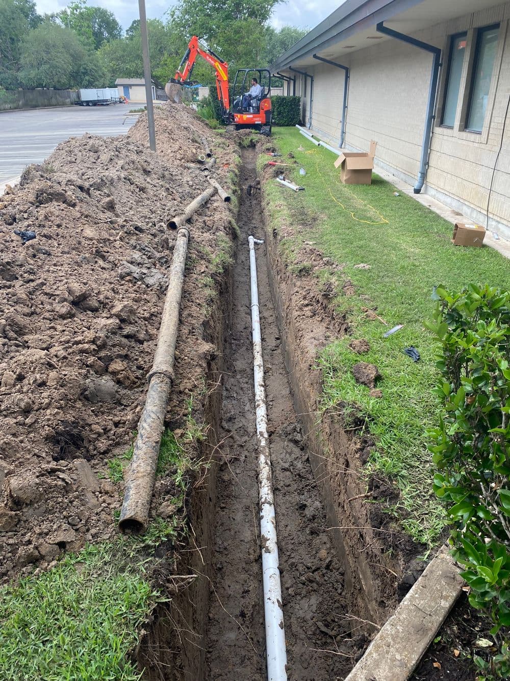 Excavation trench with pipes for construction, machinery in background, grassy area nearby.