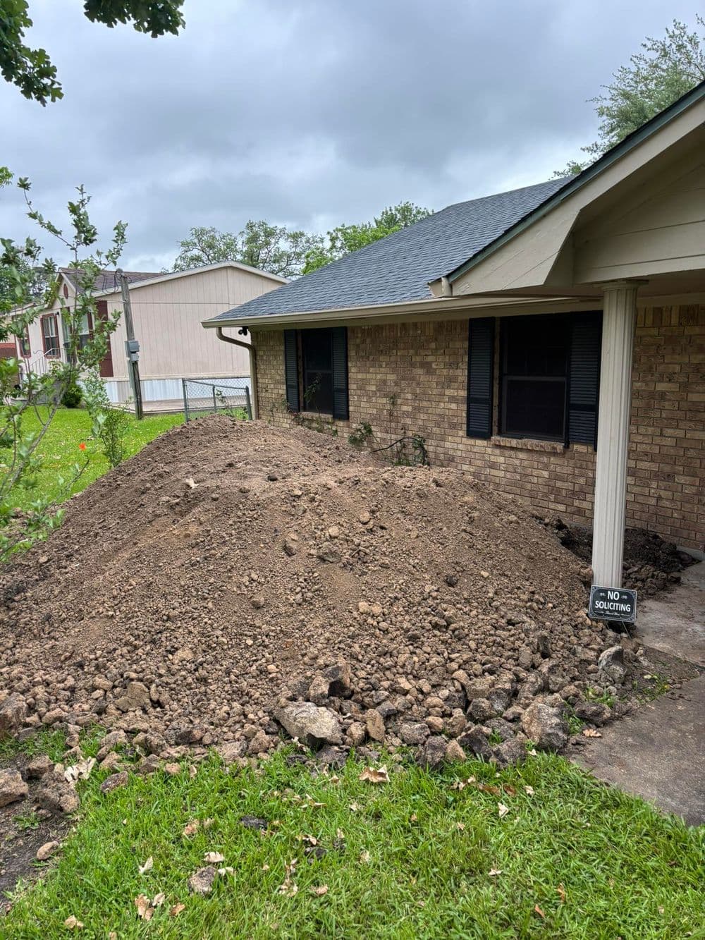 Dirt pile near house foundation, cloudy sky, residential yard with grass and trees.
