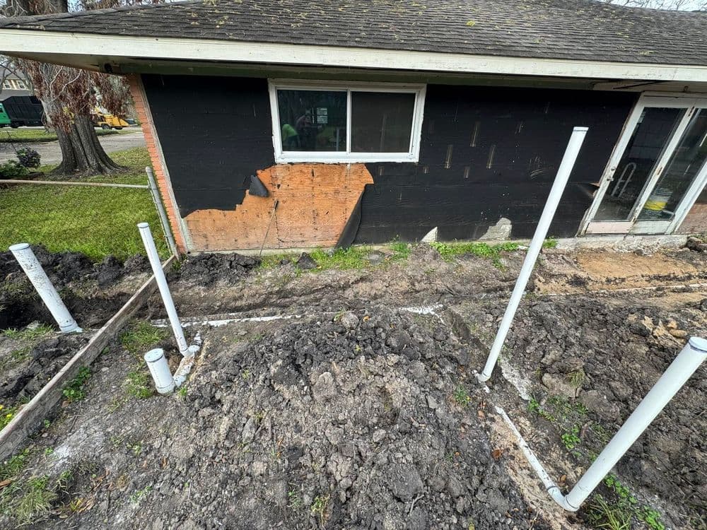 Residential construction site showing excavated area with PVC pipes near a damaged wall.